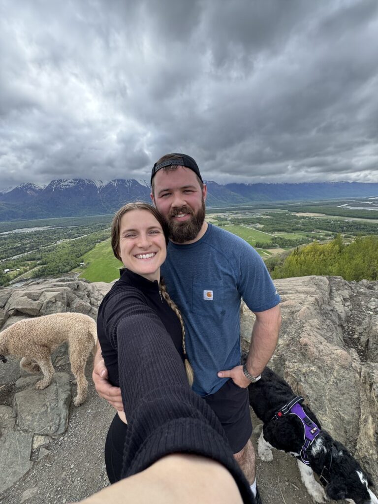A woman and man smile for a selfie atop a rocky overlook on one of their favorite Alaska hikes, with two dogs nearby and a green valley and mountains in the background under cloudy skies.