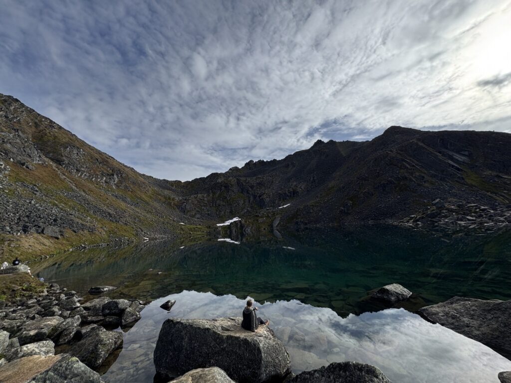 A person sits on a rock by a clear mountain lake surrounded by rocky hills under a partly cloudy sky, capturing the tranquil beauty often found on Alaska hikes.