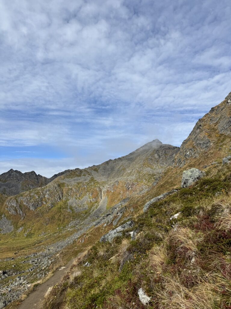 Mountain landscape with rocky slopes, grassy patches, and a dirt trail under a partly cloudy sky—perfect for those seeking classic Alaska hikes.