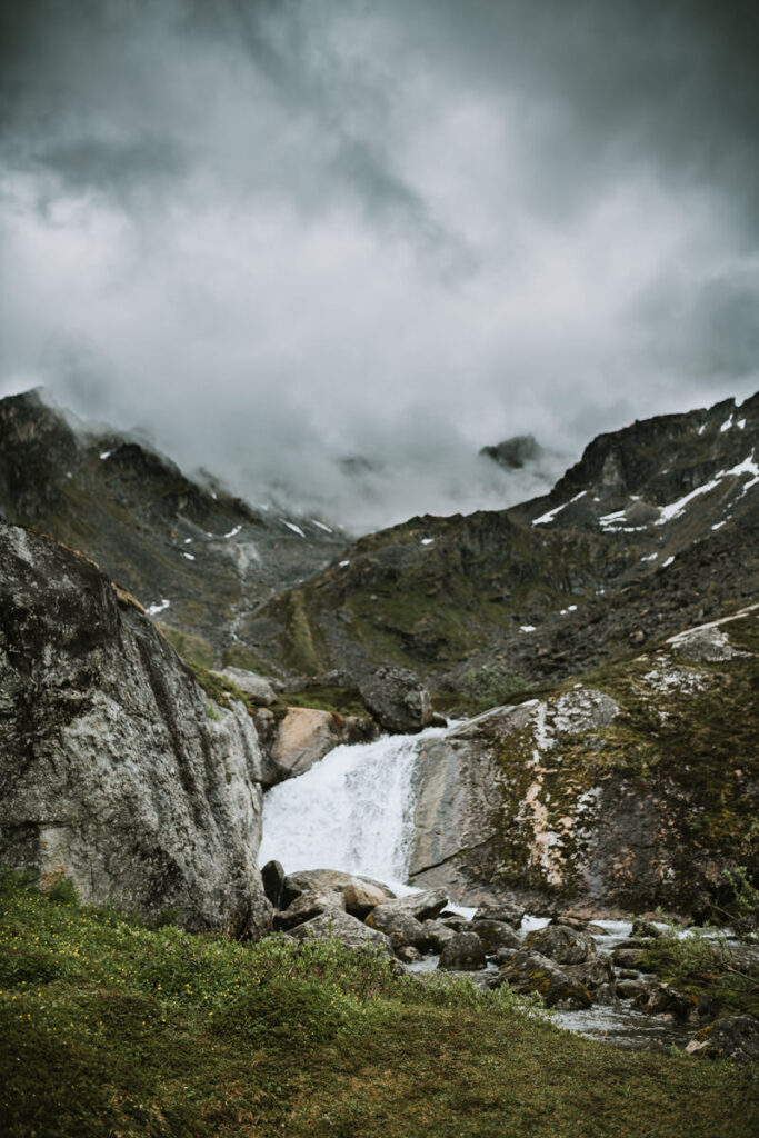 A small waterfall flows between rocky mountains under a cloudy, overcast sky, with patches of snow and green grass in the foreground—a scene reminiscent of scenic Alaska hikes.