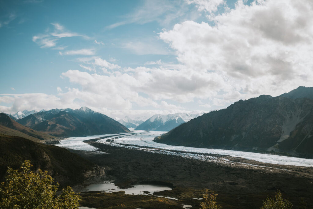 A wide river or glacier flows between two mountain ranges under a partly cloudy sky, with green vegetation and small ponds in the foreground—an iconic scene often spotted on Alaska hikes.