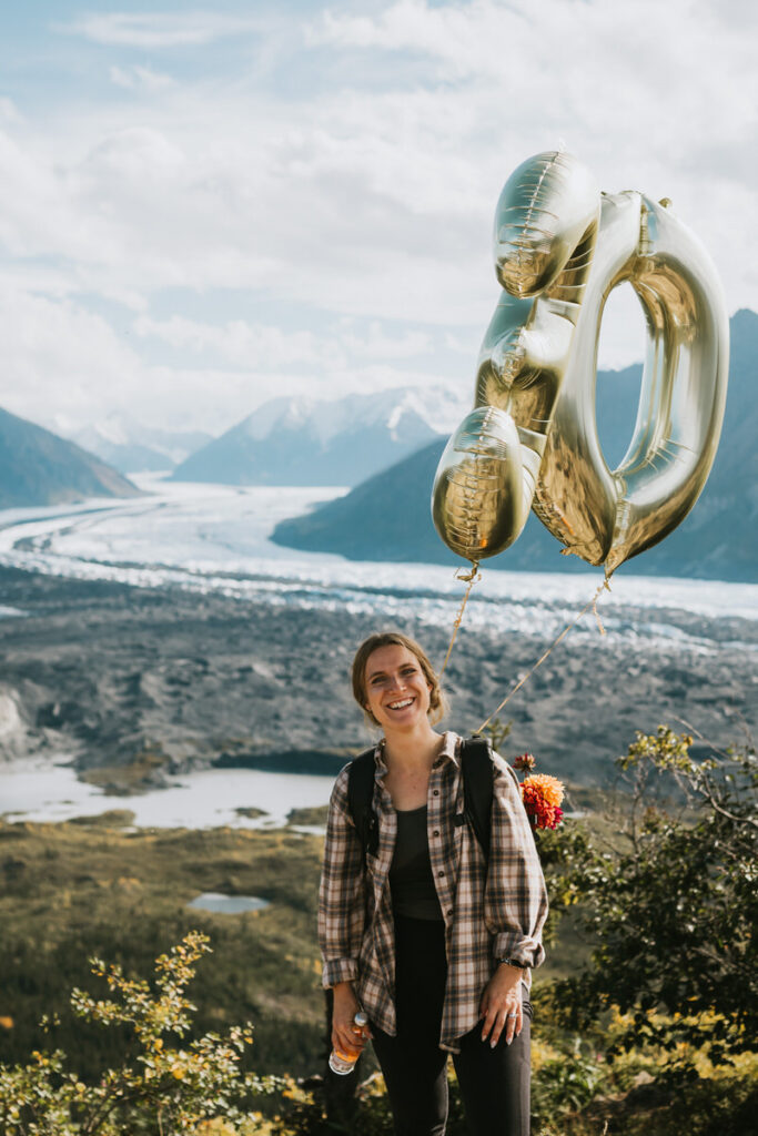 A woman with a backpack stands outdoors holding gold balloons shaped like the number 30, celebrating amidst scenic mountains and a river—an unforgettable moment inspired by classic Alaska hikes.