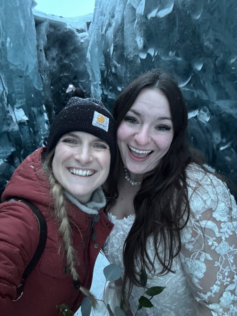 Two women smiling for a selfie in front of icy blue glacier walls after one of their favorite Alaska hikes; one wears a red jacket and beanie, the other a white lace dress.