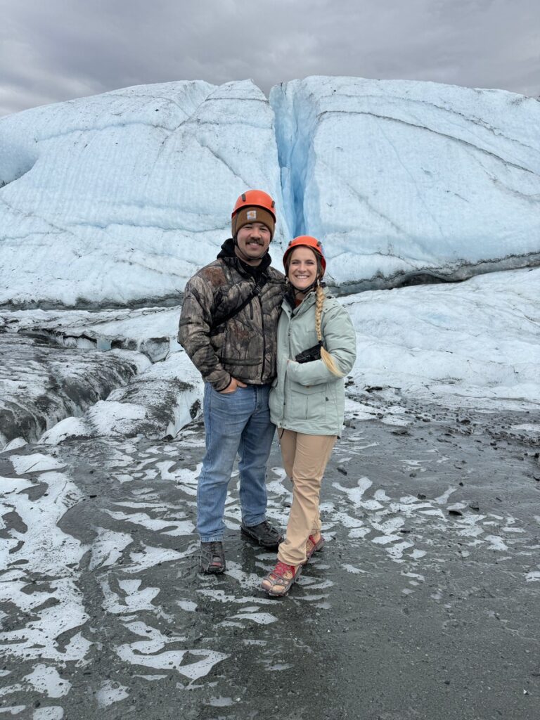 Two people wearing helmets and outdoor clothing stand together on icy ground in front of a glacier, ready to experience one of the thrilling Alaska hikes.