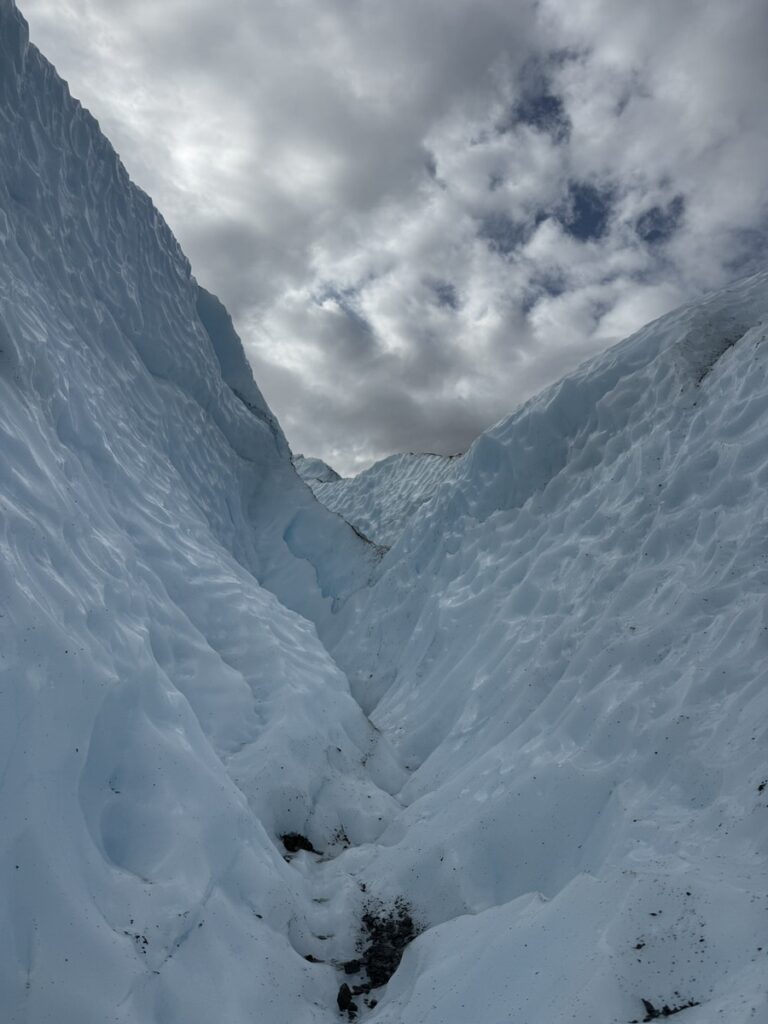 Deep crevasse in a glacier under a cloudy sky, with jagged blue-white ice walls rising on both sides—an unforgettable sight often found on Alaska hikes.
