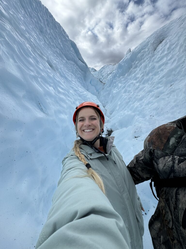 A person wearing a helmet and winter coat smiles while standing between tall walls of blue ice in a glacier crevasse, capturing the thrill of Alaska hikes.