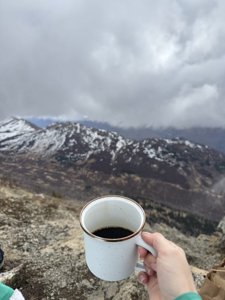 A hand holds a white mug of coffee in the foreground, with snow-capped mountains and a cloudy sky—perfect scenery for Alaska hikes—in the background.