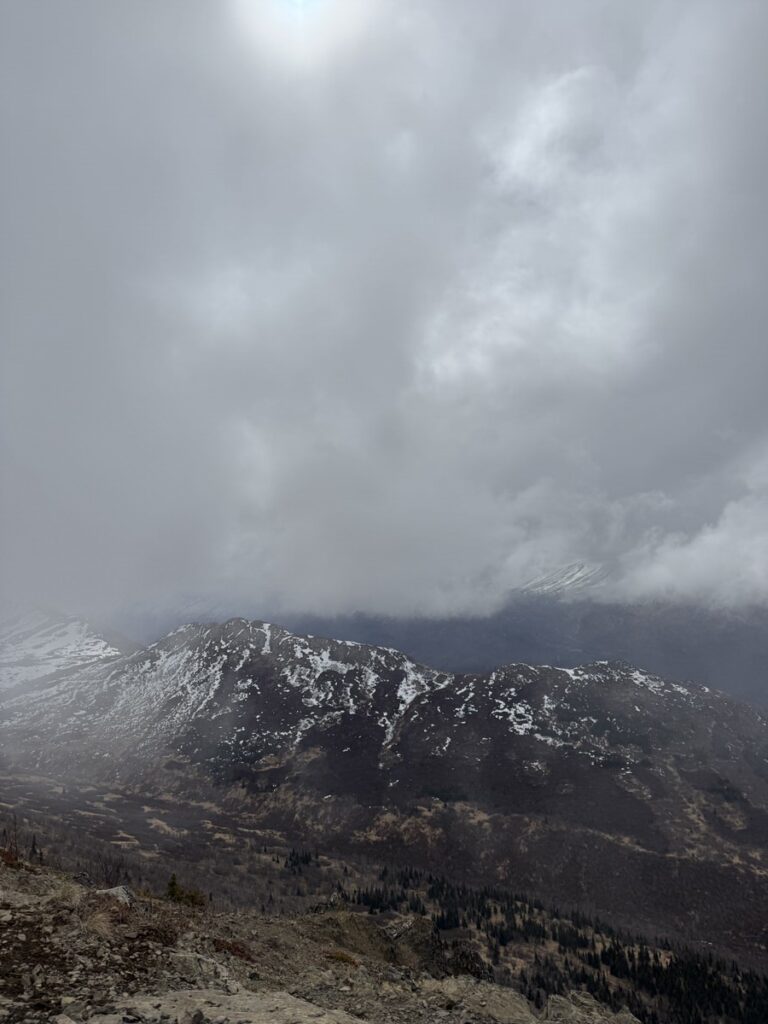 Cloudy sky over a mountain range with patches of snow on the slopes and rocky terrain in the foreground, capturing the rugged beauty often seen on Alaska hikes.