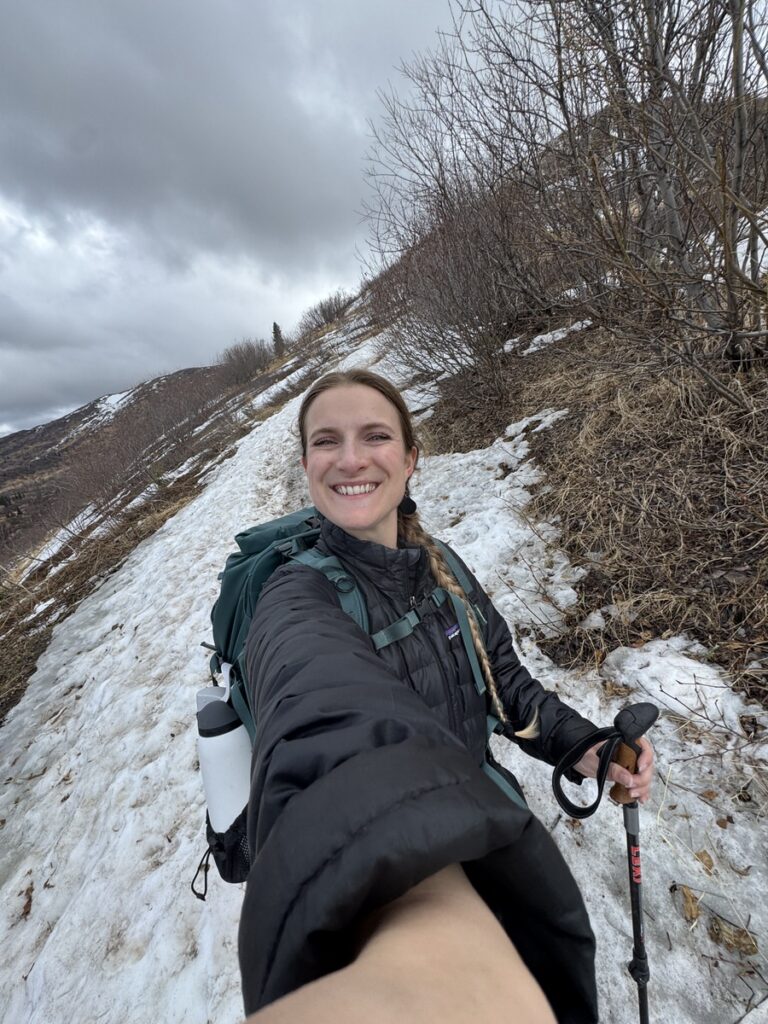 A woman hiking on a snowy mountain trail in Alaska smiles at the camera, holding a trekking pole and wearing a backpack under a cloudy sky—a perfect day for alaska hikes.