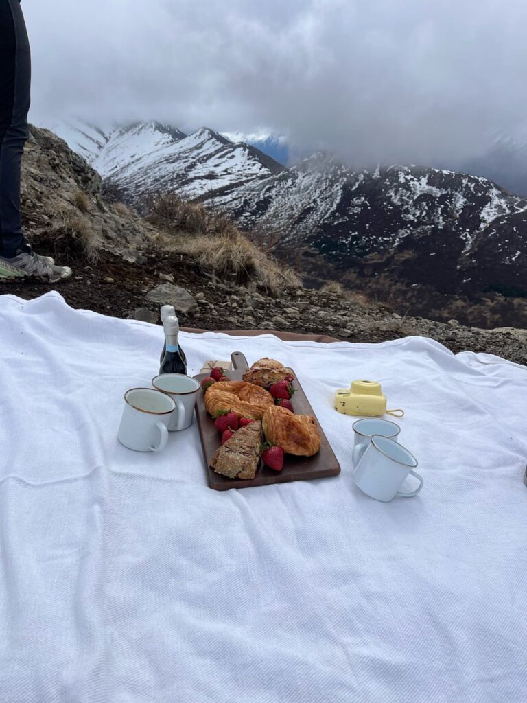 A picnic setup with pastries, mugs, a bottle, and camera on a white blanket rests on a mountainside, snowy peaks in the background—a perfect pause during Alaska hikes.