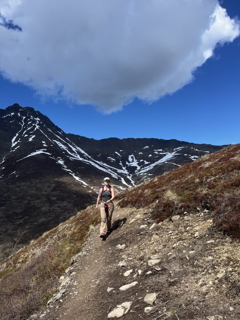 A person wearing hiking gear walks along a rocky mountain trail under a large cloud, with snow-capped peaks in the background—an iconic scene from alaska hikes.