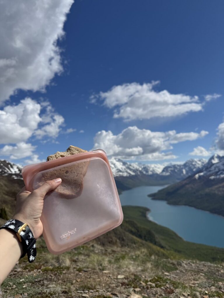 A hand holds a sandwich in a reusable silicone bag during one of many Alaska hikes, with a mountain lake and snow-capped peaks in the background under a partly cloudy sky.