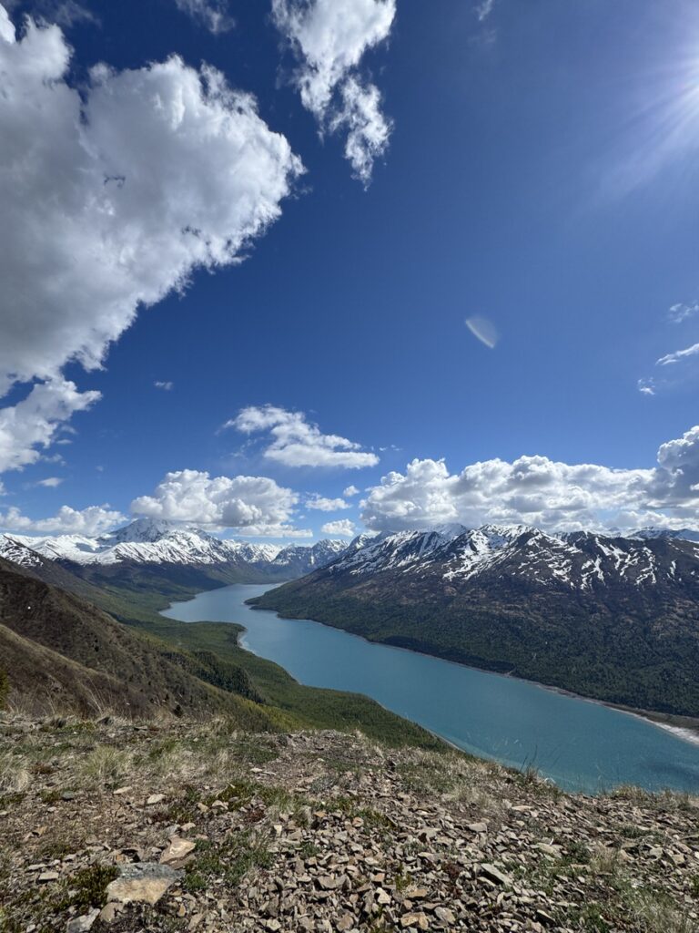 A clear river winds through a valley with snow-capped mountains under a blue sky with scattered clouds—an inspiring scene often found on Alaska hikes, with rocky ground visible in the foreground.