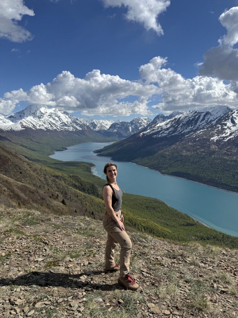 A person stands on a rocky hillside overlooking a blue river winding through a valley with snow-capped mountains—an iconic scene from Alaska hikes—under a partly cloudy sky.