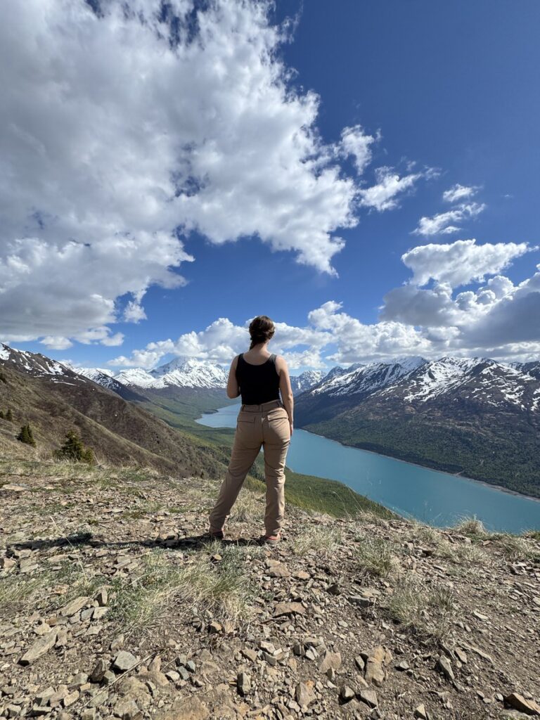 A person stands on a rocky hilltop overlooking a blue lake, surrounded by snow-capped mountains under a partly cloudy sky—a breathtaking scene often found on Alaska hikes.