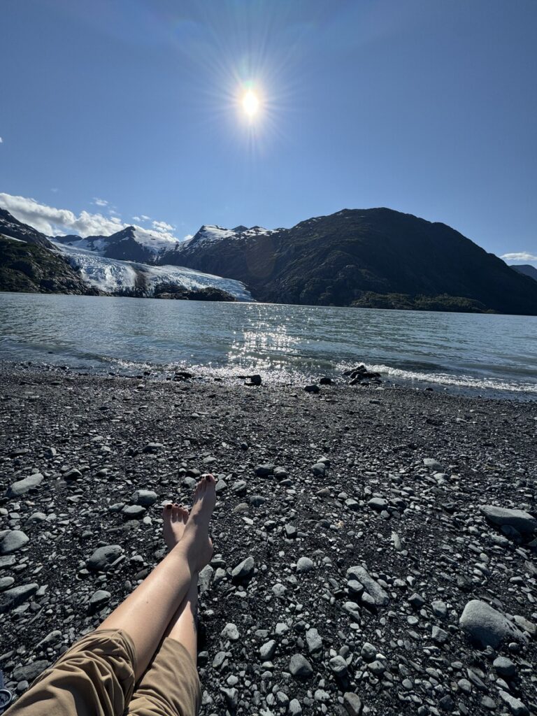 Person sits on a rocky shore with legs outstretched, facing a lake, glacier, and mountains under a sunny, clear blue sky—capturing the serene beauty found on classic Alaska hikes.