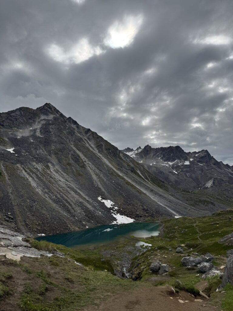 Overcast sky above a mountain range with rocky slopes, patches of snow, and a small turquoise lake nestled in the valley below—an inspiring scene often found on Alaska hikes.