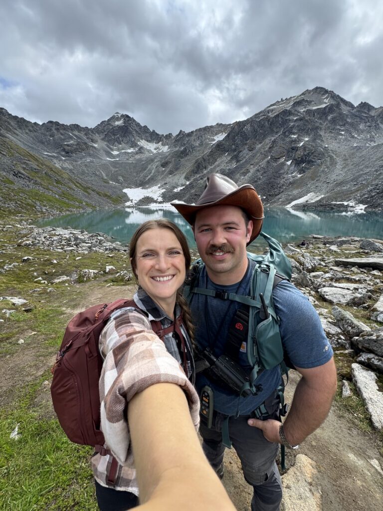 Two hikers take a selfie in front of a mountain lake, surrounded by rocky terrain and snow-capped peaks under a cloudy sky—capturing the wild beauty of Alaska hikes.