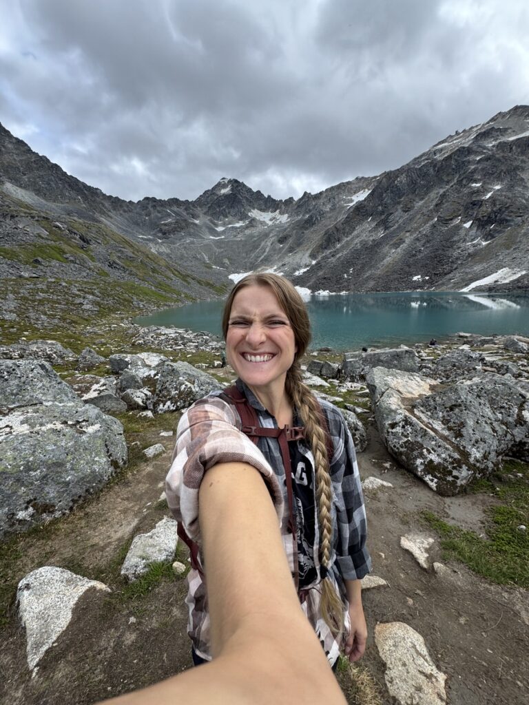 A person taking a selfie while smiling in front of a turquoise alpine lake surrounded by rocky mountains under a cloudy sky, capturing the beauty of Alaska hikes.