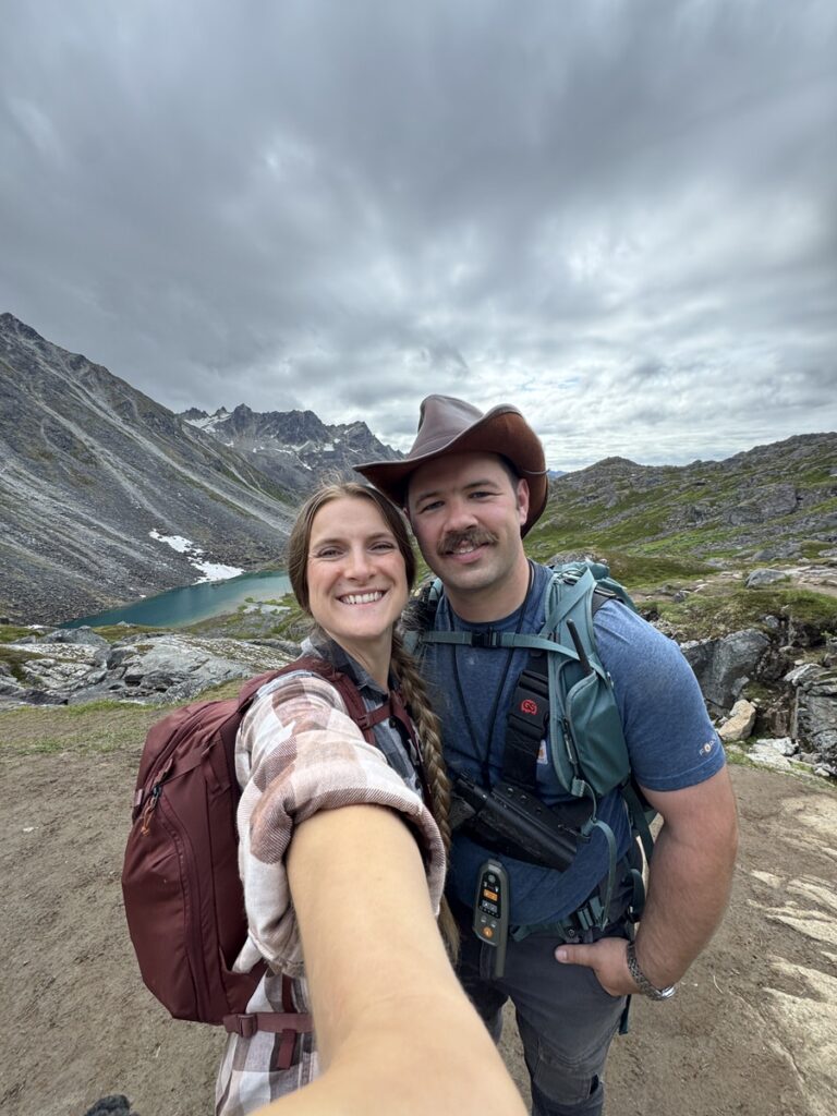Two people taking a selfie while hiking on an Alaska trail, with a lake and rugged peaks in the background under a cloudy sky—capturing the spirit of unforgettable Alaska hikes.