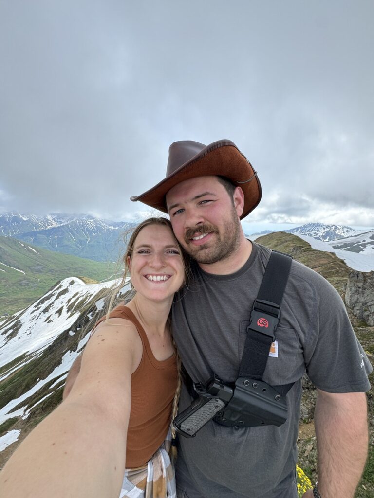 A smiling couple poses for a selfie on a snowy mountain during one of their Alaska hikes, with cloudy skies and distant peaks in the background. The man wears a cowboy hat and the woman wears a tank top.