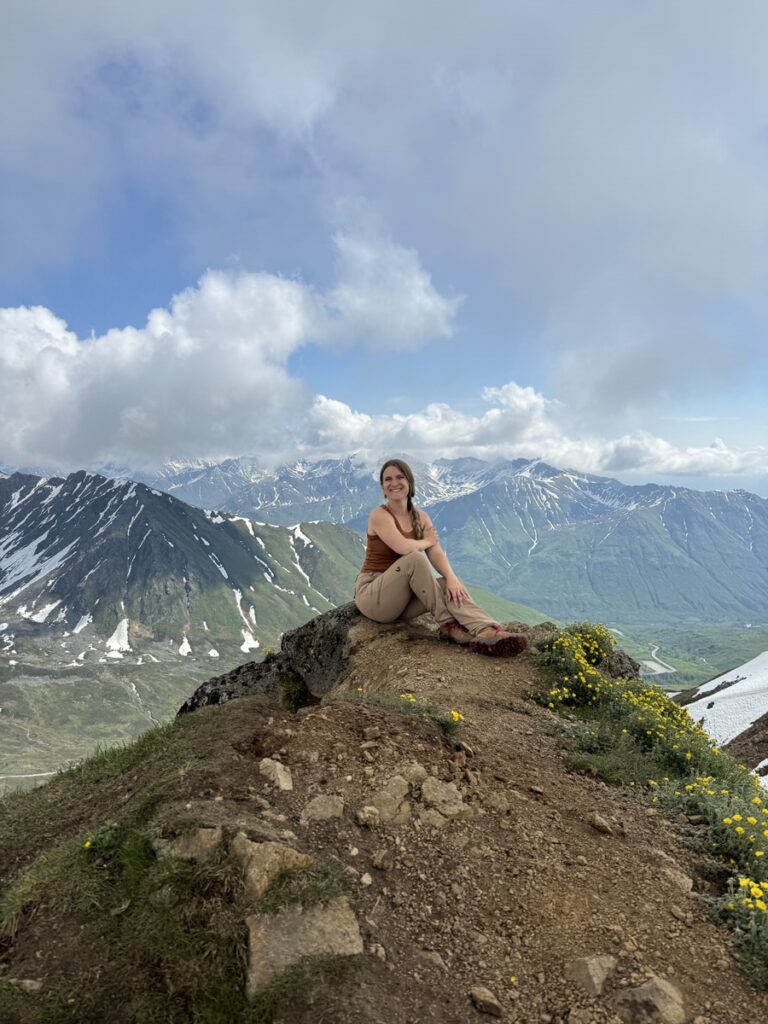 A woman sits on a rocky hilltop during one of her Alaska hikes, with mountains, patches of snow, and clouds in the background. Yellow wildflowers grow nearby.