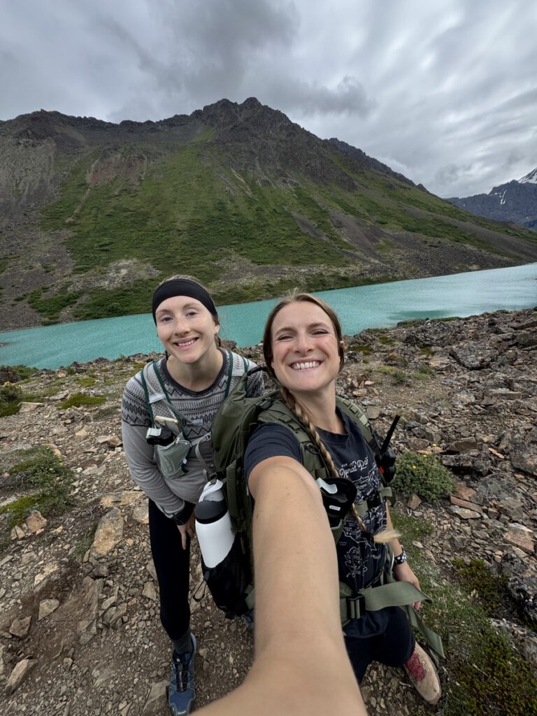 Two women in hiking gear smile for a selfie by a turquoise lake along one of the scenic Alaska hikes, with rocky terrain and green mountains in the background under a cloudy sky.