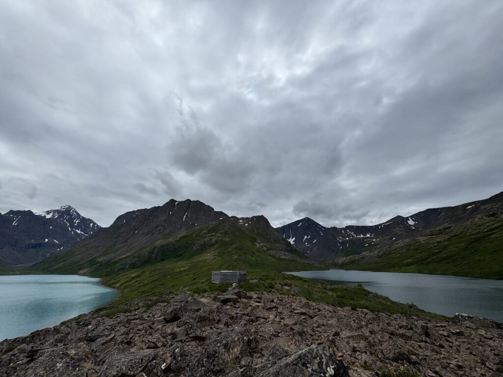 Rocky foreground leads to a small, rectangular structure; two lakes and green mountains sit beneath a cloudy sky in the background, evoking the rugged beauty found on alaska hikes.
