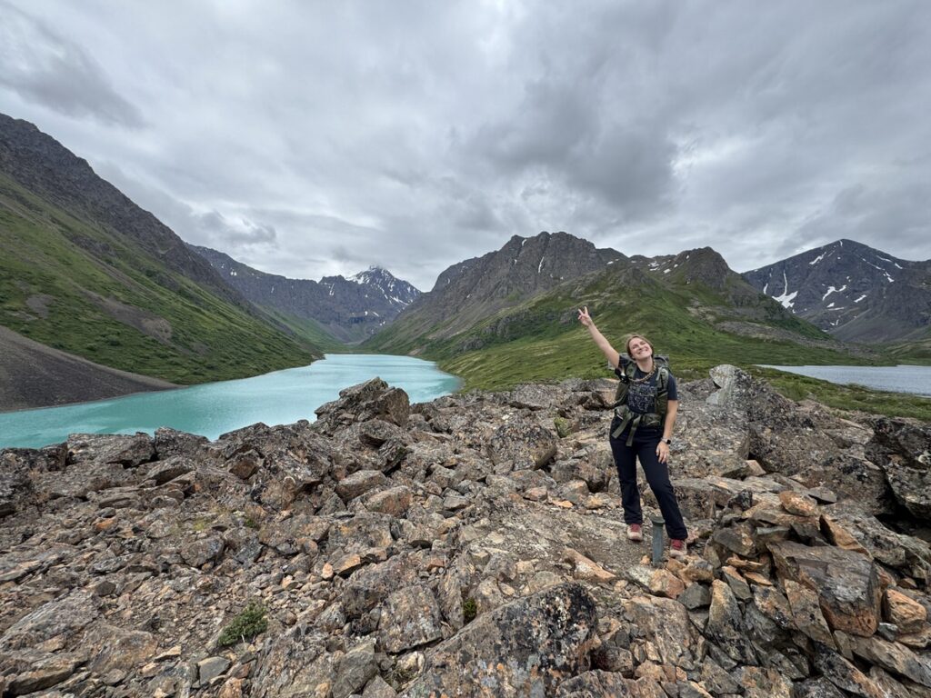 Person standing on rocky terrain, pointing towards a turquoise lake and mountains under a cloudy sky—an awe-inspiring scene reminiscent of challenging Alaska hikes.