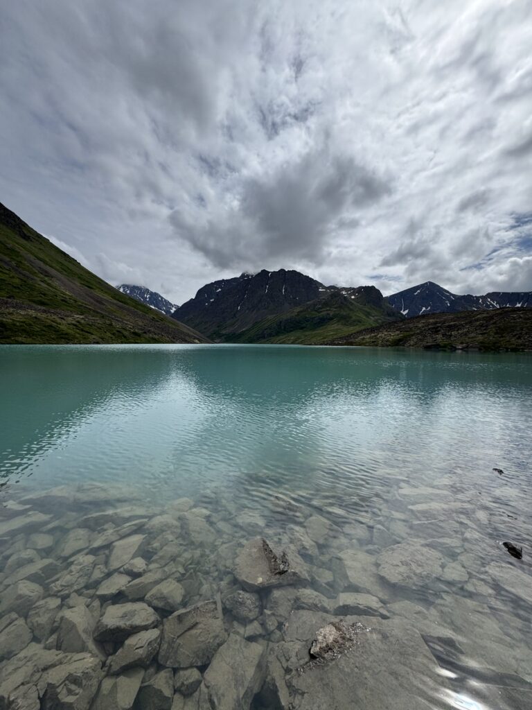 A turquoise lake with clear water and a rocky shoreline, set against green hills and snow-capped mountains under a cloudy sky—perfect scenery often found on unforgettable Alaska hikes.