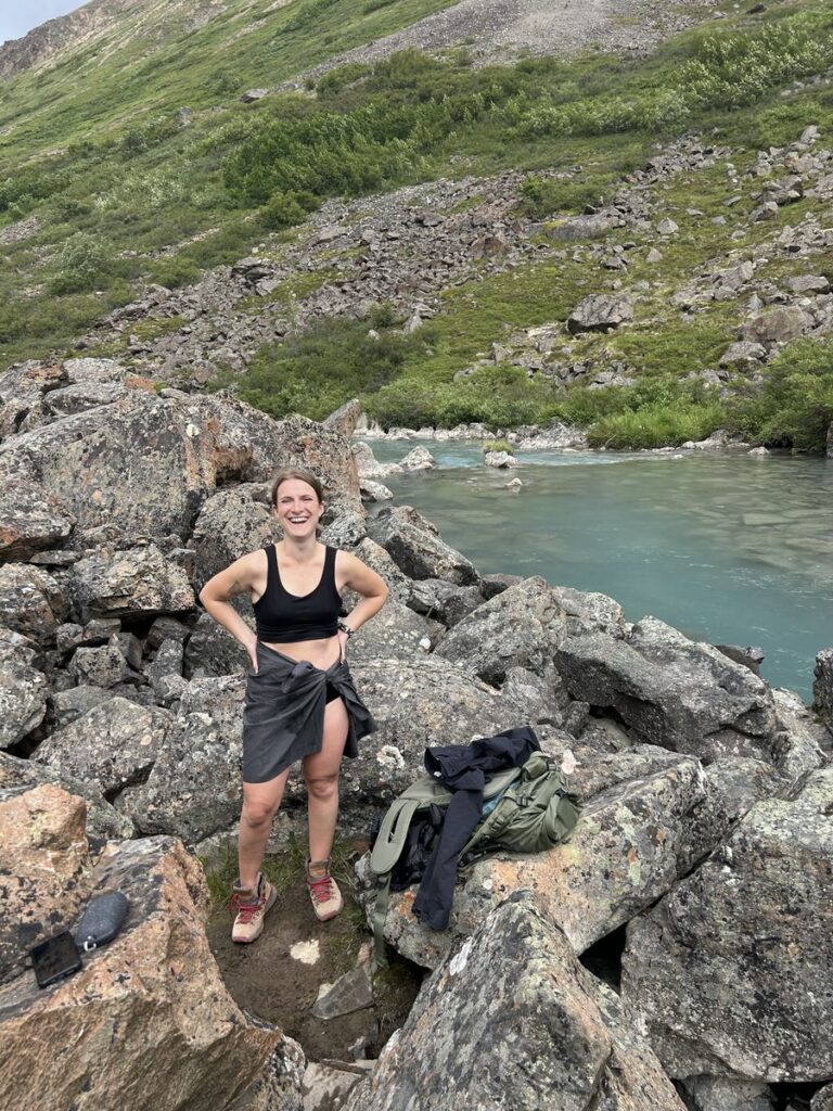 A person in outdoor clothing stands on rocky terrain beside a turquoise river, surrounded by green hills and vegetation—an inspiring scene reminiscent of classic Alaska hikes. Outdoor gear is placed on the rocks nearby.