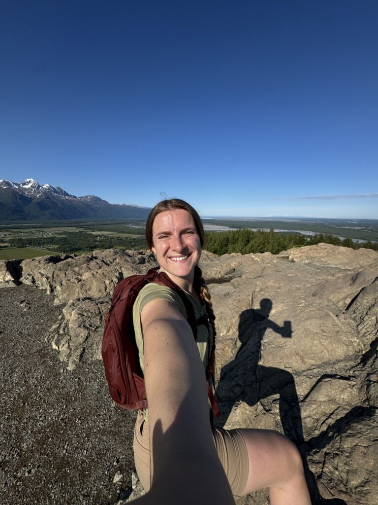A woman with a braid and a backpack takes a selfie on a rocky outcrop amid stunning mountains and trees, capturing the spirit of Alaska hikes under a clear blue sky.