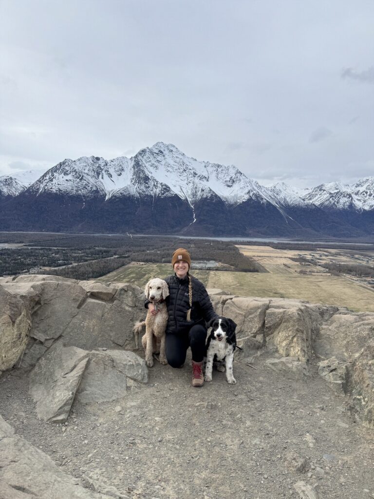 A person wearing a brown hat kneels on a rocky ledge with two dogs, taking in the stunning views of snowy mountains and a valley—an unforgettable moment amidst classic Alaska hikes.