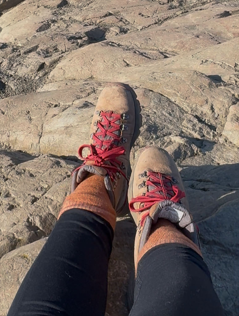 A person wearing tan hiking boots with red laces, orange socks, and black pants sits on rocky terrain in sunlight, ready to explore rugged Alaska hikes.