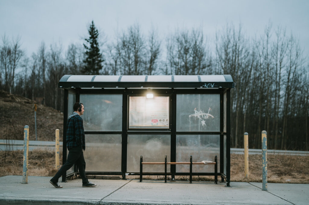 A man walks past an empty bus shelter on a cloudy day, with bare trees and a road in the background.
