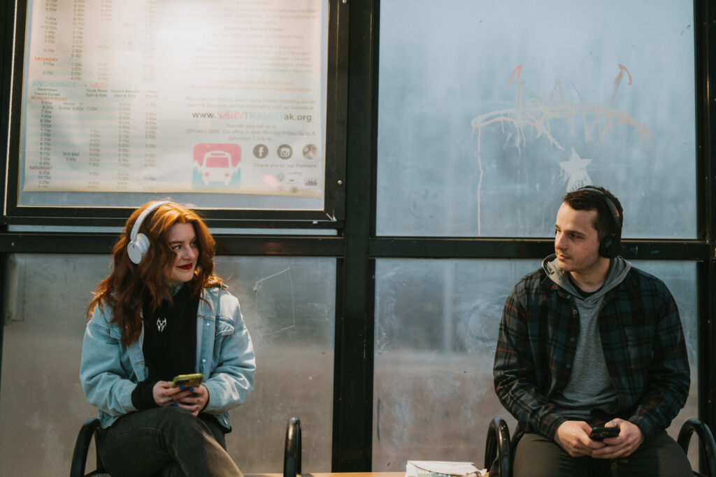 Two people wearing headphones sit apart on a bench at a bus stop, each holding a phone, with a schedule visible behind them.