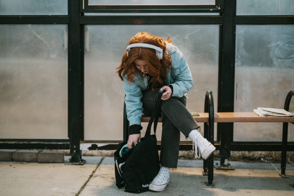 A person with red hair wearing headphones sits on a bench at a bus stop, reaching into a black backpack on the ground beside them.