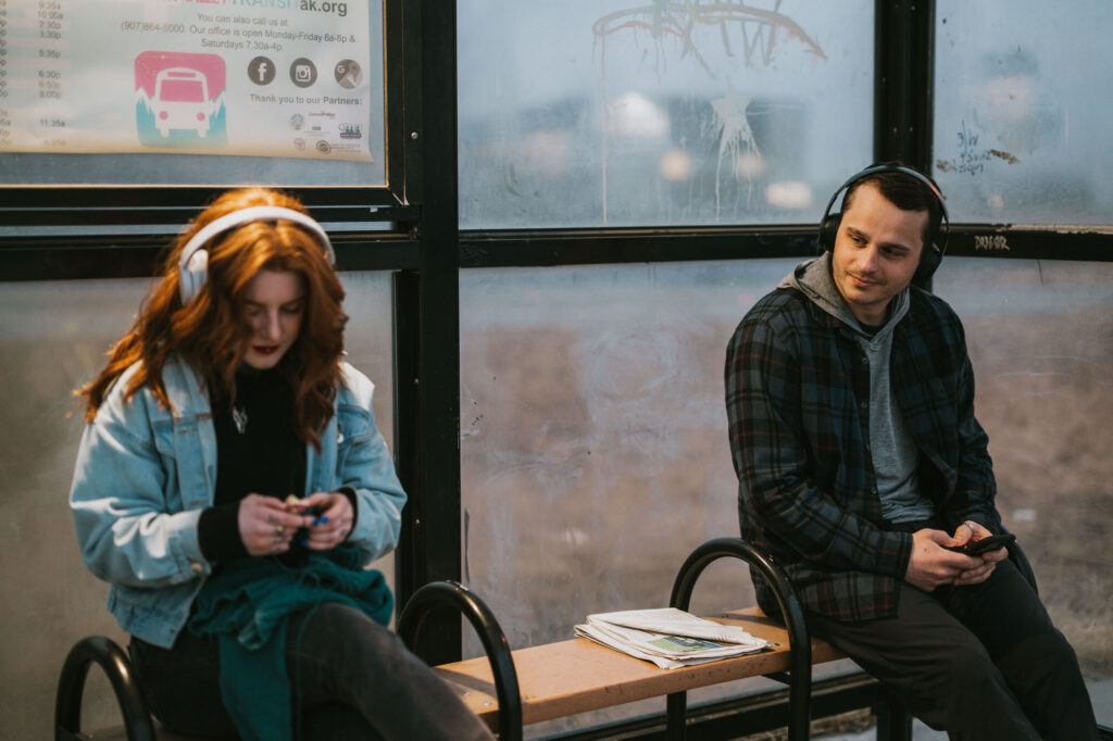 Two people wearing headphones sit on a bench at a bus stop. The woman looks at her phone, while the man looks at her and holds his phone. A stack of newspapers is beside them.