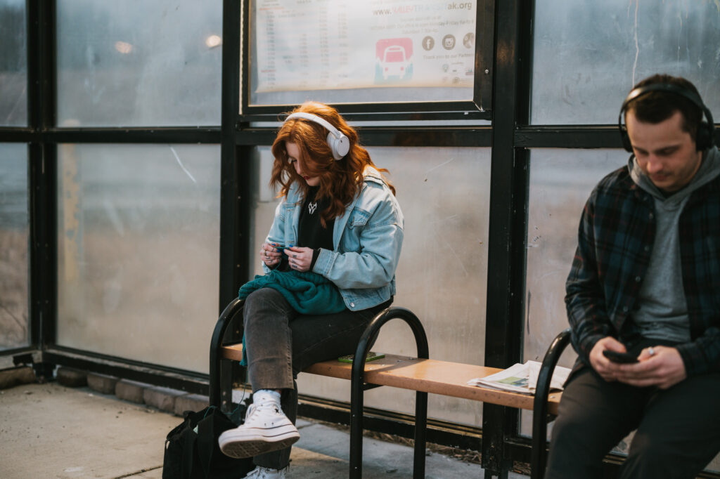 Two people sit on a bench at a bus stop; both are wearing headphones. The woman on the left is knitting, and the man on the right is looking at his phone.