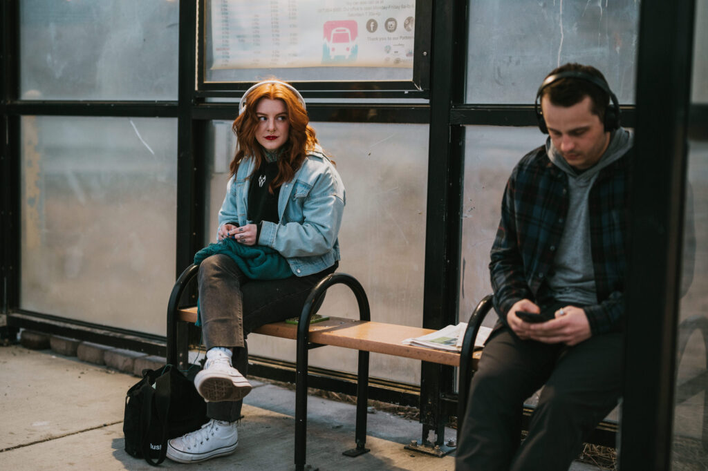 Two people sit on a bench at a bus stop; one woman looks ahead while a man with headphones looks at his phone.