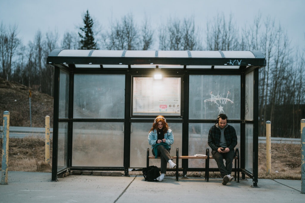 Two people sit apart on a bench at a bus stop shelter, both looking at their phones. The shelter is surrounded by bare trees and overcast skies.