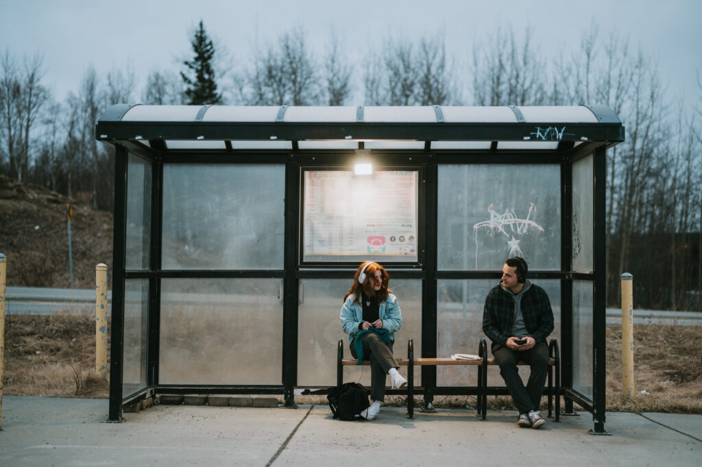 Two people sit on a bench at a covered bus stop, facing each other, with leafless trees and a road in the background.