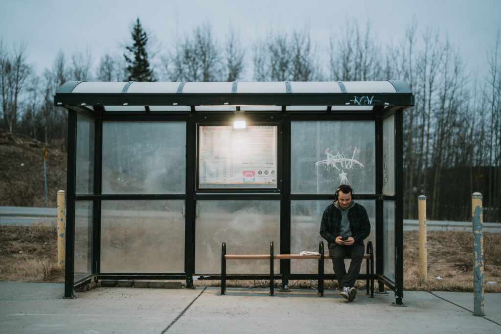 A person sits alone on a bench at a bus stop, looking at their phone. The shelter is glass with graffiti, and trees without leaves are visible in the background.