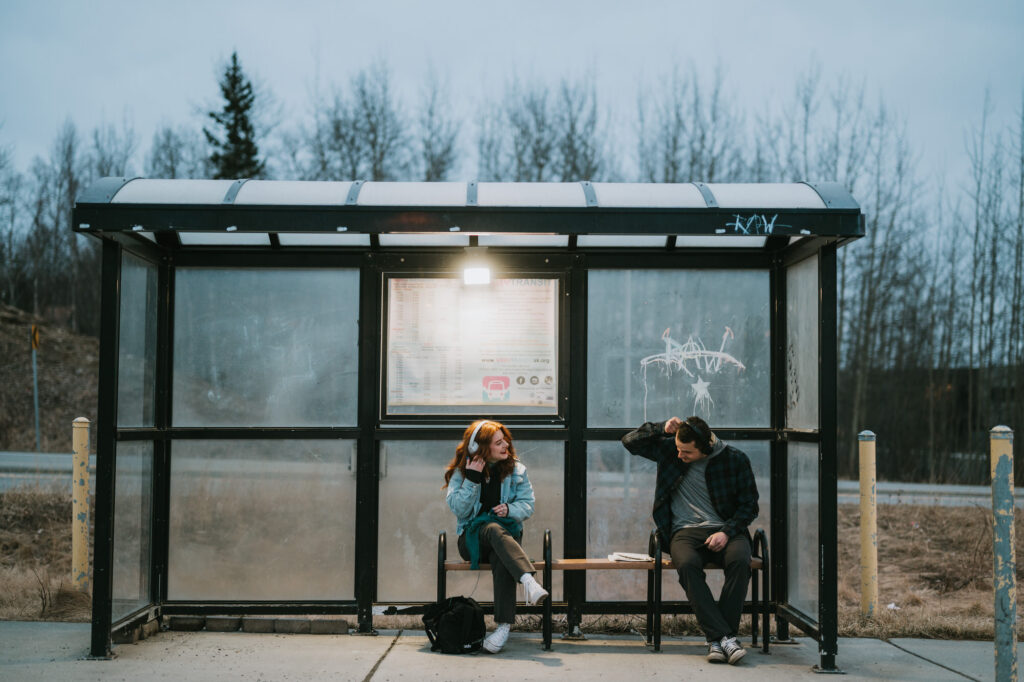 Two people sit on benches at a bus stop shelter, talking. It is cloudy and the surrounding trees are bare. One person has a backpack on the ground.