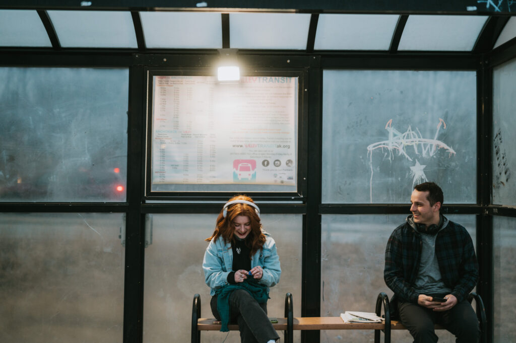 Two people sit on a bench at a bus stop; one is listening to headphones and looking down, while the other looks over at them. A transit schedule is posted behind them.