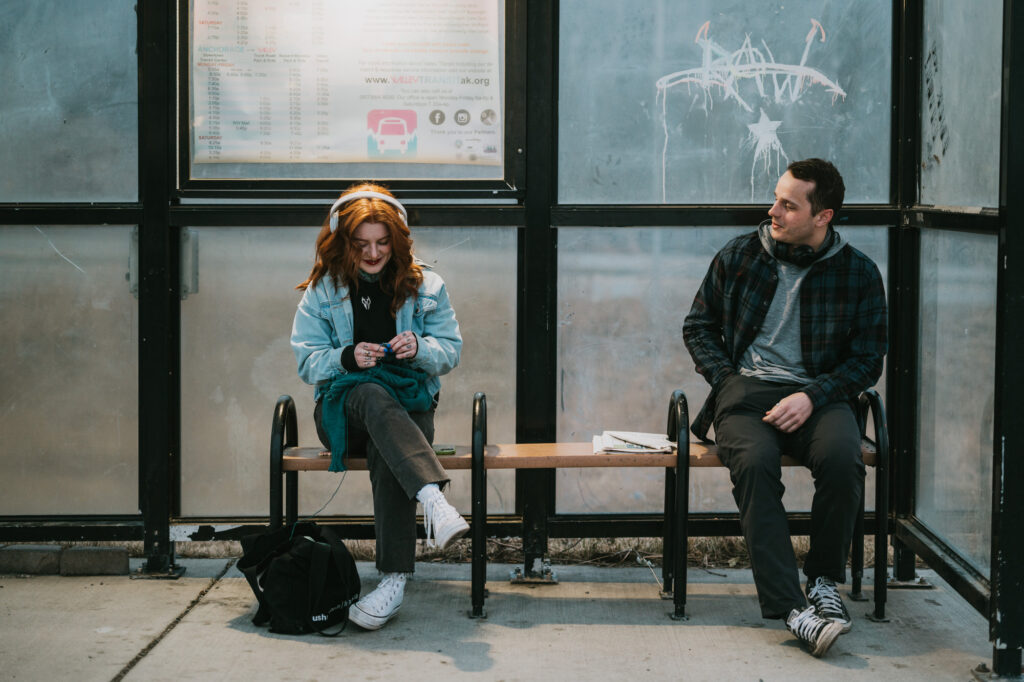 A woman with headphones sits on a bench at a bus stop, looking at her phone, while a man sits a few feet away, looking at her and smiling.