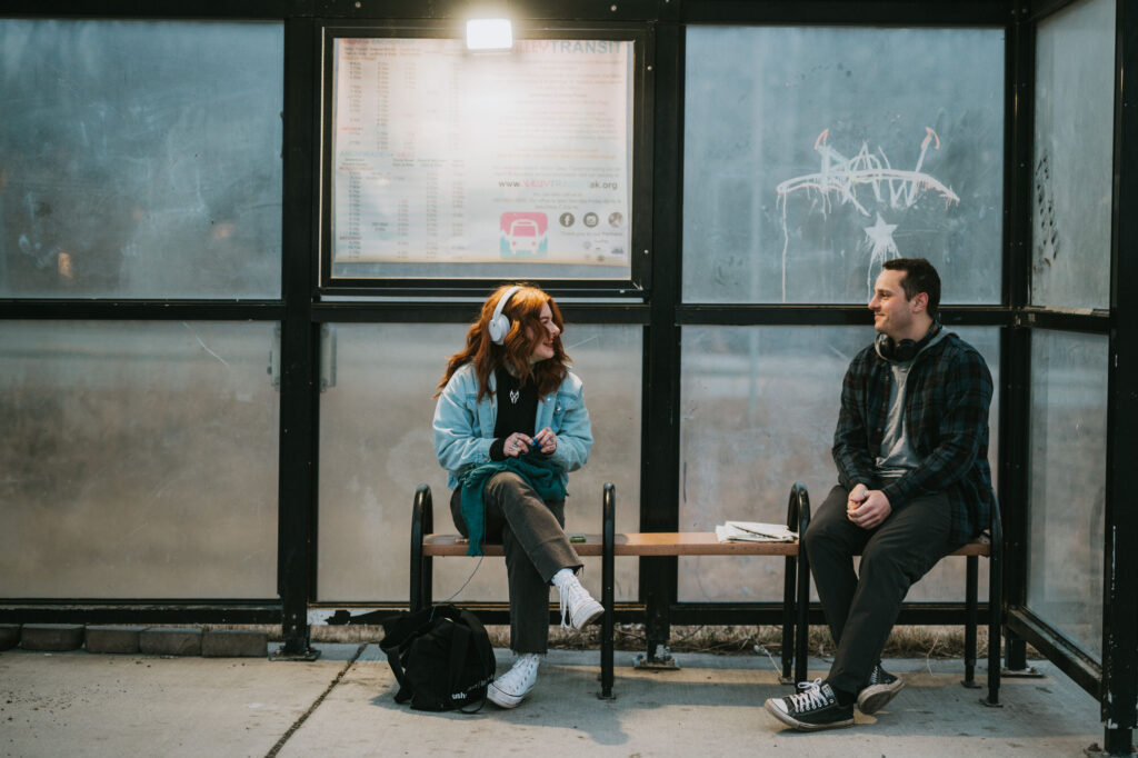 Two people sit on a bench at a bus stop, wearing headphones and facing each other. A backpack and a notebook are on the ground and bench between them.