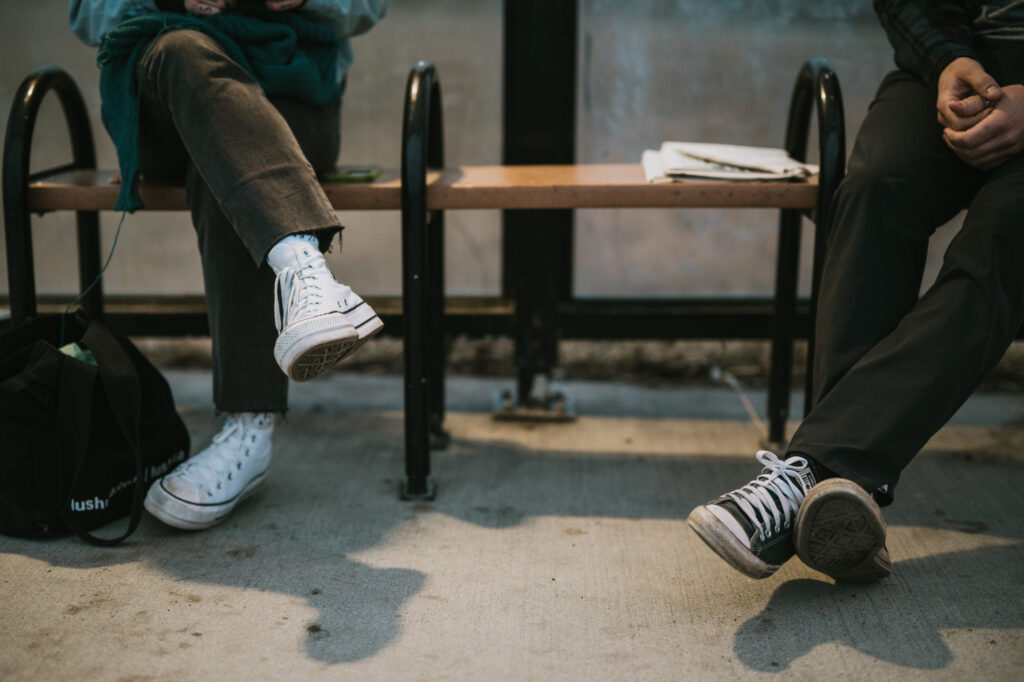 Two people sit on a bench, legs crossed, wearing casual clothes and Converse sneakers. Only their lower bodies are visible. A black bag sits on the ground beside them.