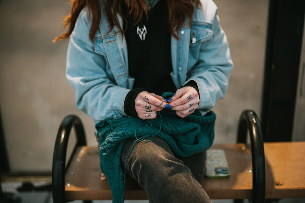 A person sits on a bench, knitting with teal yarn. They wear a denim jacket, dark jeans, and have tattoos on their fingers. A phone rests on the bench beside them.