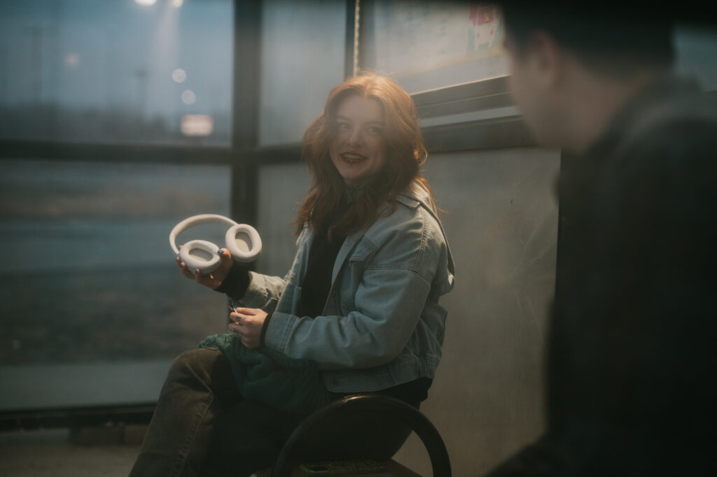 A person with long red hair sitting indoors holds white headphones while looking at another person who is mostly out of focus.